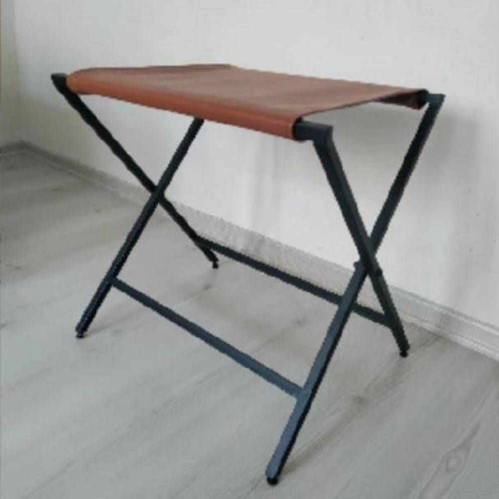 A foldable metal suitcase rack with a genuine leather top displayed in a minimalist interior setting on a wooden floor