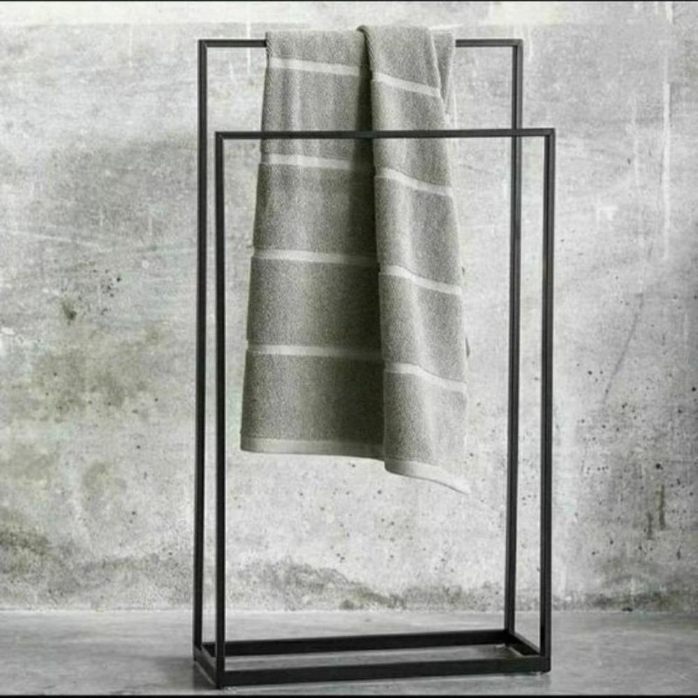 Modern free-standing black metal towel holder displayed against a textured gray background, with a neatly folded towel hanging from the top bar, showcasing minimalist design and functionality for contemporary bathroom decor.