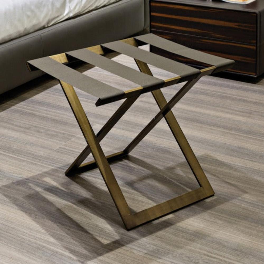 A close-up of a foldable gold metal luggage rack with leather straps, positioned on a sleek wooden floor near a modern bedside table.