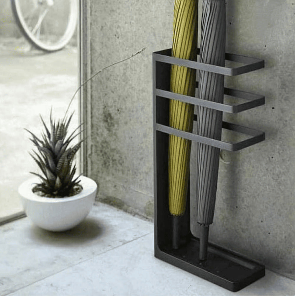 A black metal umbrella stand holding two umbrellas, displayed near a window with a modern concrete backdrop and a decorative plant 