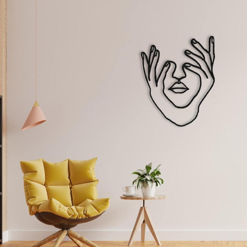 Minimalist metal wall art of a woman's face with hands framing it, mounted on a neutral wall above a cozy setup with a mustard-yellow cushioned chair, a wooden side table, and a small plant.