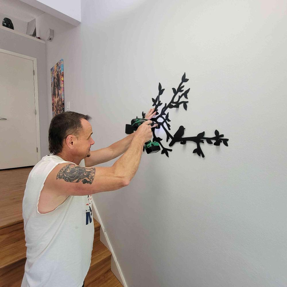 A man is using a drill to install a black metal wall-mounted coat rack with bird and branch designs. The rack is being secured to the wall in a modern entryway, adding a functional and decorative element to the space.