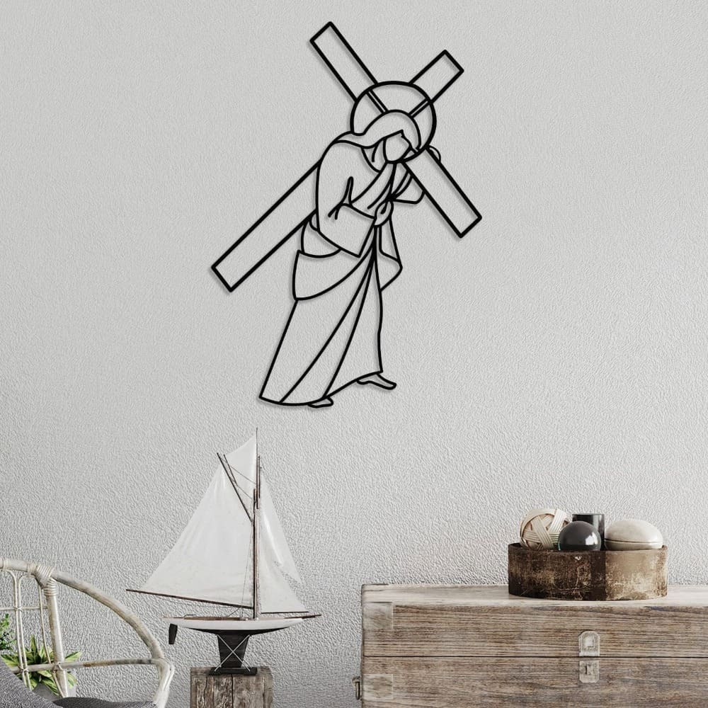 Black metal wall art depicting Jesus carrying the cross, mounted on a light gray textured wall. Below, a rustic wooden chest and a wicker chair sit alongside a decorative sailboat and a bowl filled with natural ornaments, adding a coastal-inspired touch.