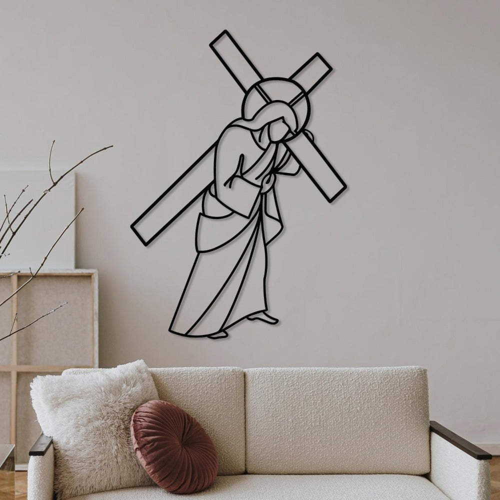 Black metal wall art depicting Jesus carrying the cross, mounted on a light beige wall in a modern living room. The room features a cream-colored sofa adorned with a fluffy white pillow and a round maroon accent pillow, complemented by minimalist wooden decor and dried branches.