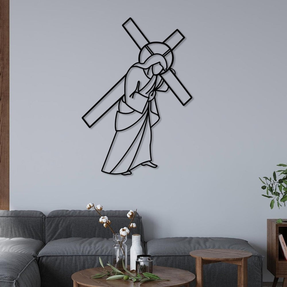 Black metal wall art depicting Jesus carrying the cross, mounted on a light gray wall in a cozy living room. Below, a modern gray sectional sofa is complemented by a wooden coffee table with vases and greenery, creating a serene and reflective space.