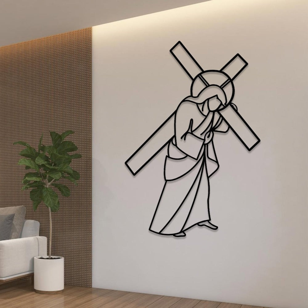 Black metal wall art depicting Jesus carrying the cross, mounted on a clean white wall with soft lighting in a modern interior. A nearby area features a white armchair and a tall potted plant, adding a touch of greenery to the peaceful setting.