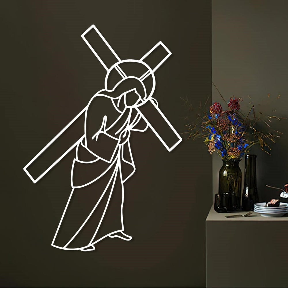 White metal wall art depicting Jesus carrying the cross, mounted on a dark wall. The setting includes a modern console table decorated with a black vase filled with colorful dried flowers and neatly stacked plates, creating a contemporary yet spiritual ambiance.