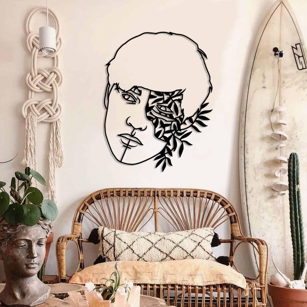 A black metal wall art piece showcasing a man's face and floral patterns, mounted on a textured wall in a beach-inspired room featuring a rattan bench and a surfboard. I