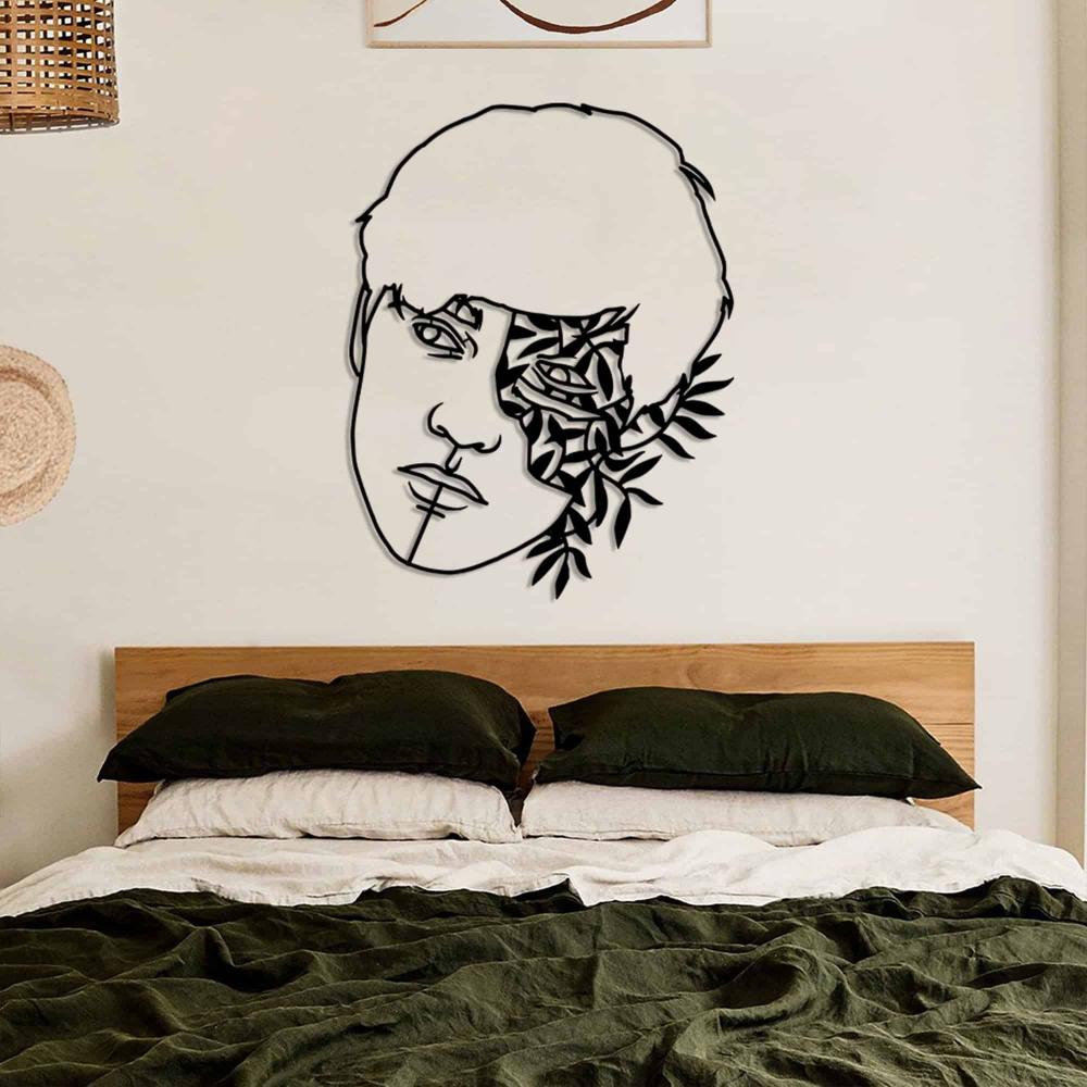 A black metal wall art piece depicting a man's face with floral and leaf motifs, mounted above a bed with dark green bedding in a minimalist bedroom.