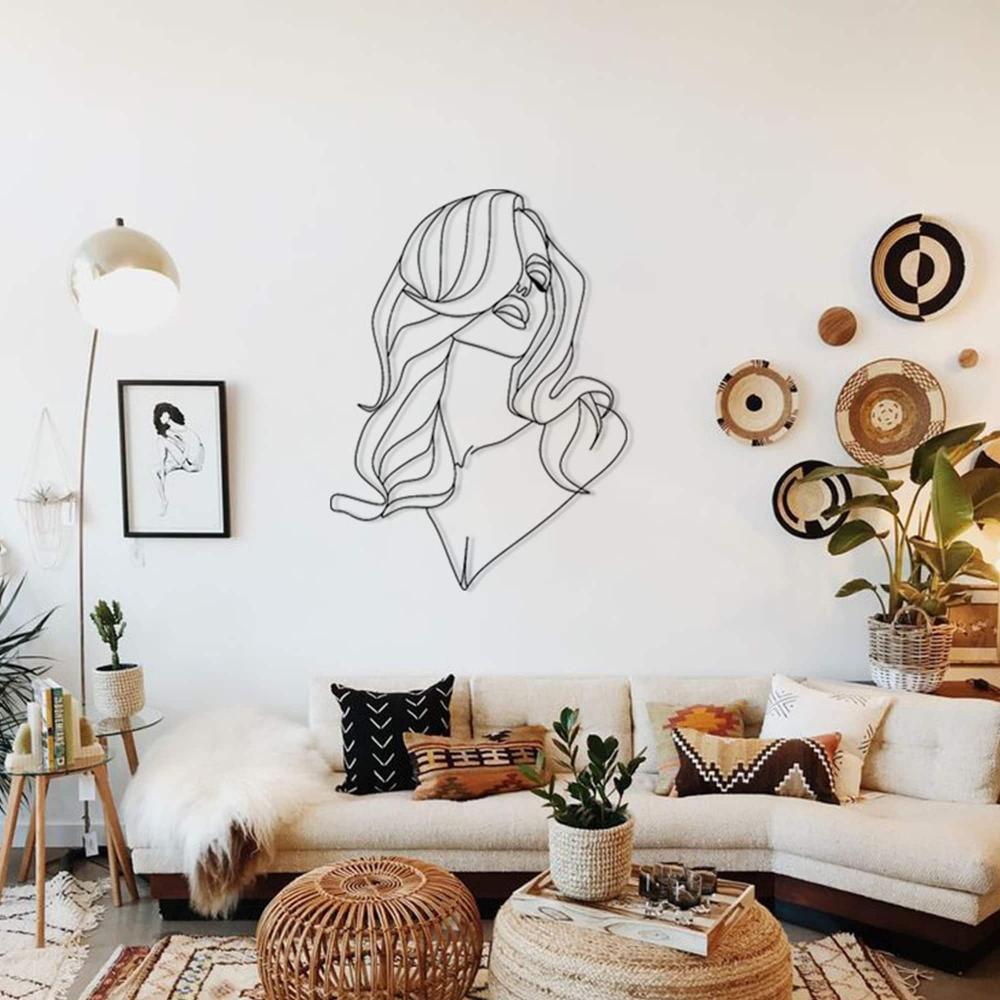 Modern line art metal wall decor of a woman with long hair mounted on a white wall in a stylish boho-chic living room. The room features a beige sectional sofa adorned with patterned throw pillows, woven baskets, indoor plants, and wall-mounted woven plates for a warm, natural aesthetic.