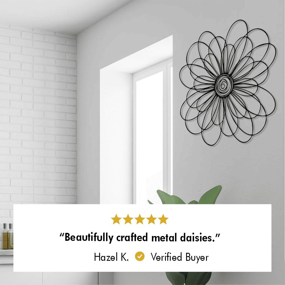 A close-up view of a metal daisy-shaped wall art mounted near a window in a bright, minimalist room. Below the artwork is a 5-star customer review from a verified buyer, Hazel K., stating: "Beautifully crafted metal daisies."