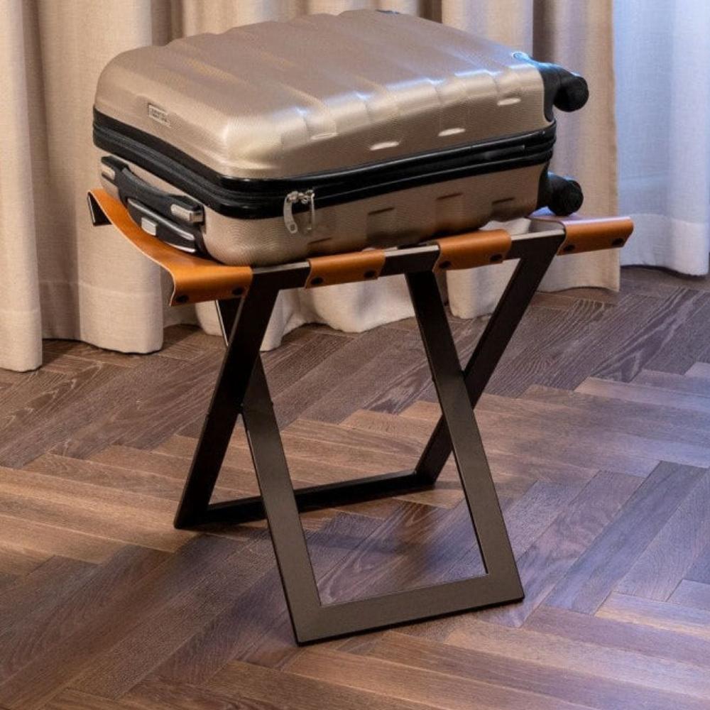 A beige suitcase resting on a modern luggage rack with a metallic frame and caramel leather straps on a wooden floor.