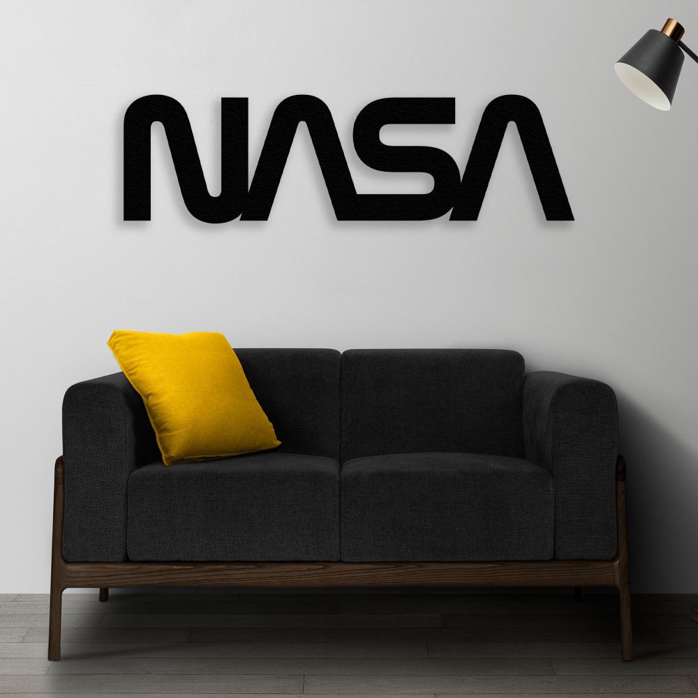 Modern living room with a black NASA metal sign wall art mounted on a white wall. Below the sign is a dark gray sofa with a yellow accent pillow, and a black wall lamp positioned to the right.