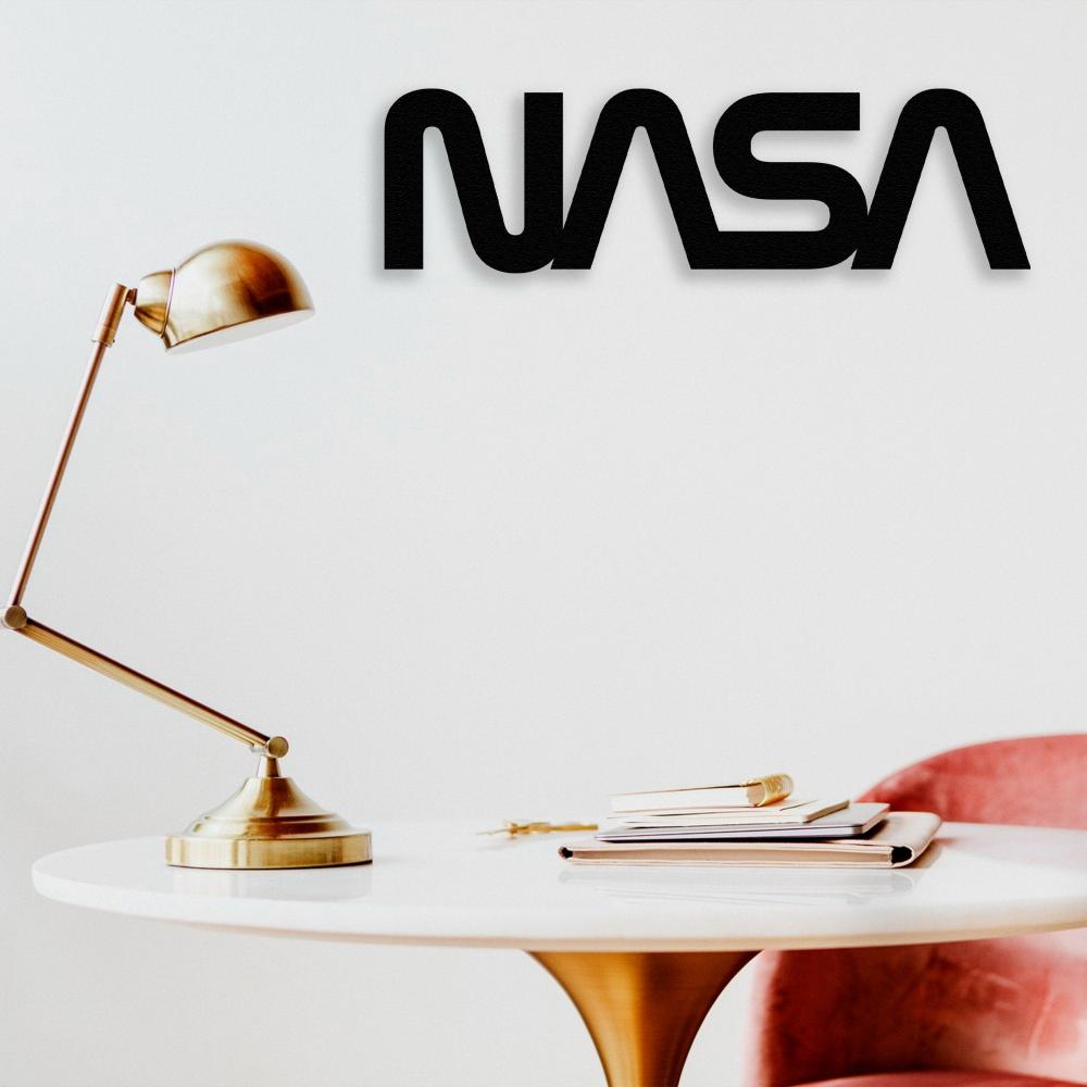 Minimalist workspace featuring a black NASA metal sign wall art mounted on a plain white wall. Below the sign is a white round desk with a gold lamp and neatly stacked notebooks, accompanied by a pink chair.