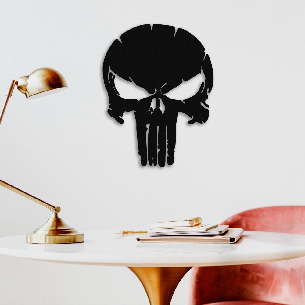 A black Punisher skull metal wall art displayed on a plain white wall above a round table with a gold lamp, books, and a red velvet chair.