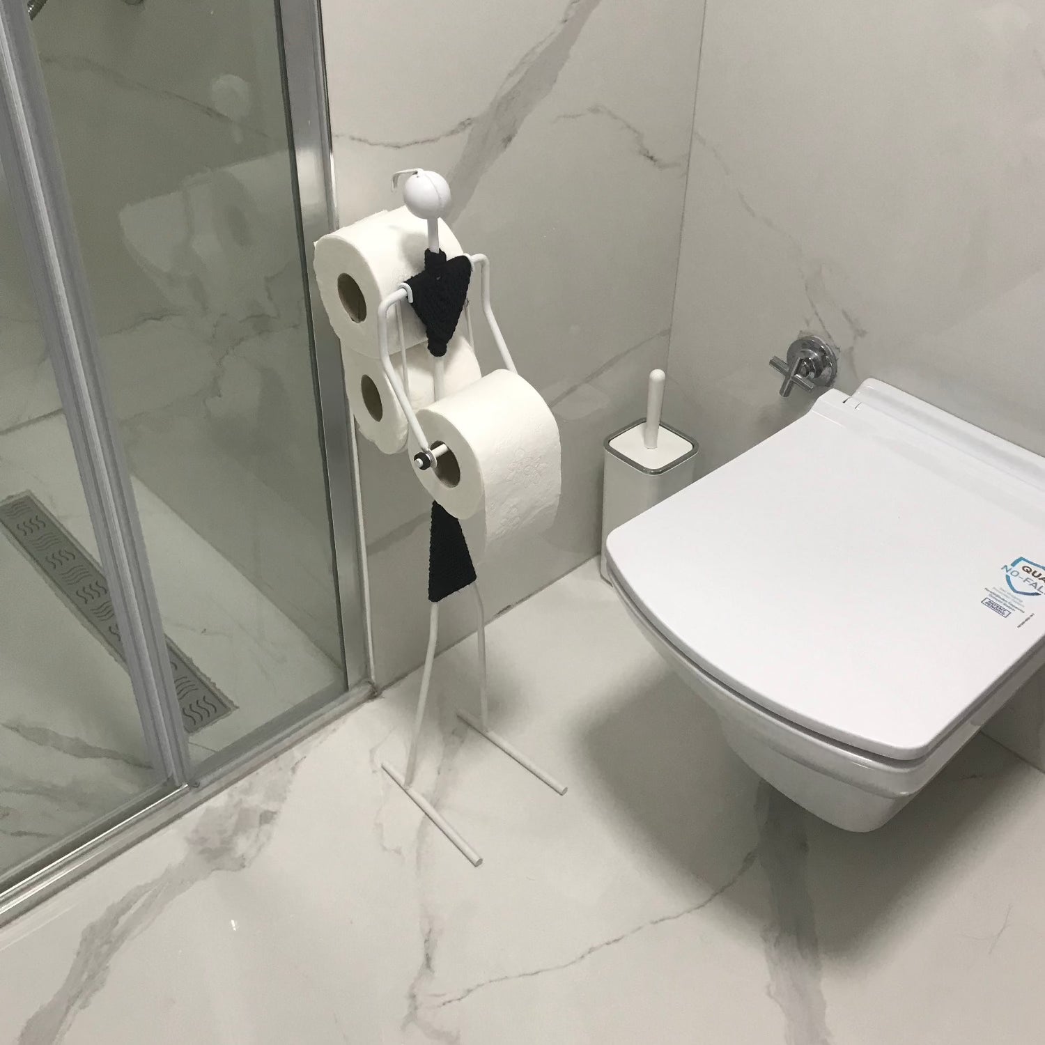 A modern bathroom with white marble flooring. A sleek, wall-mounted toilet is complemented by a white minimalist toilet paper holder that holds several rolls. The shower area is enclosed with glass doors.