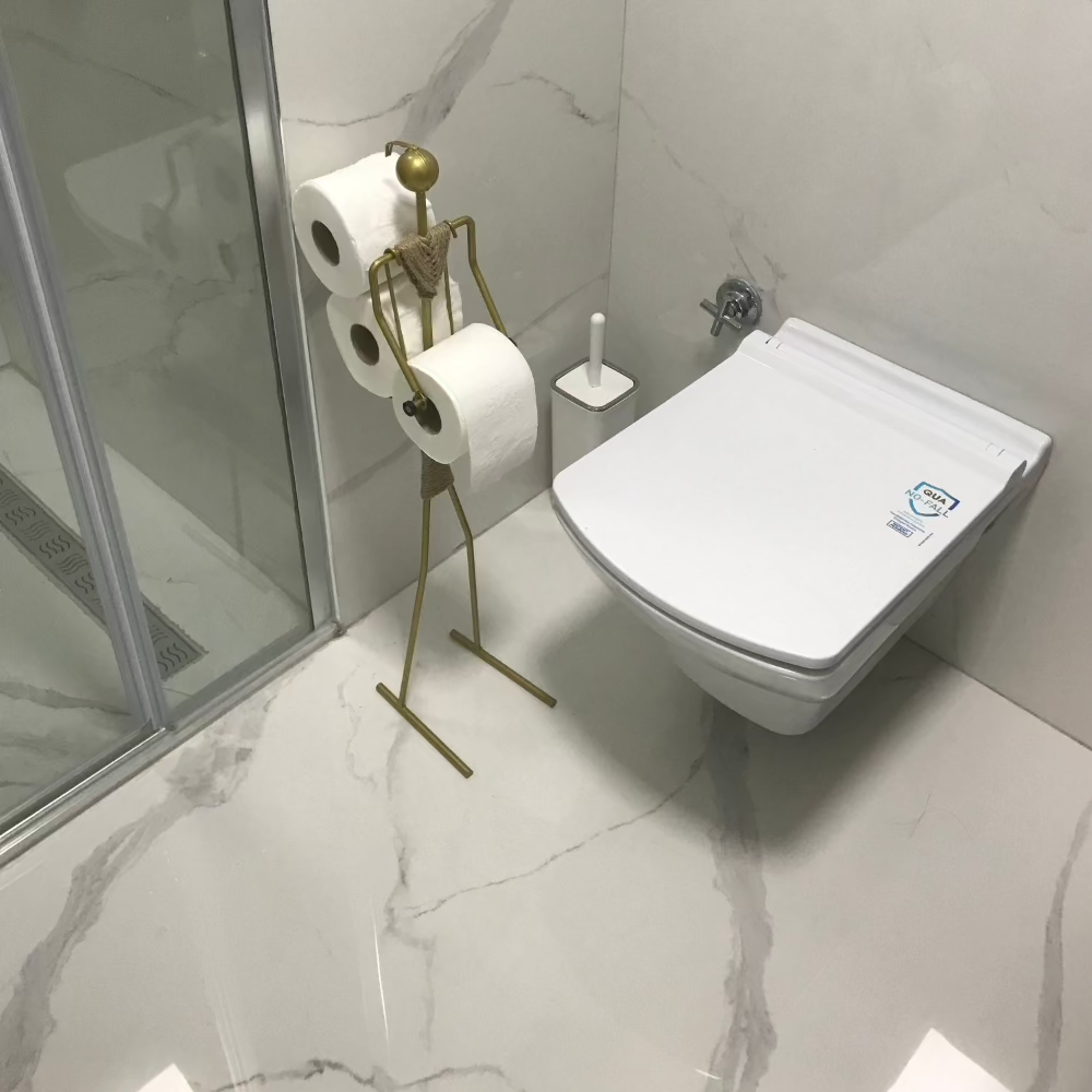 A modern bathroom featuring a sleek, wall-mounted toilet next to a stylish, minimalist toilet paper holder in gold, holding multiple rolls of toilet paper. The bathroom has a luxurious marble finish and a glass-enclosed shower.