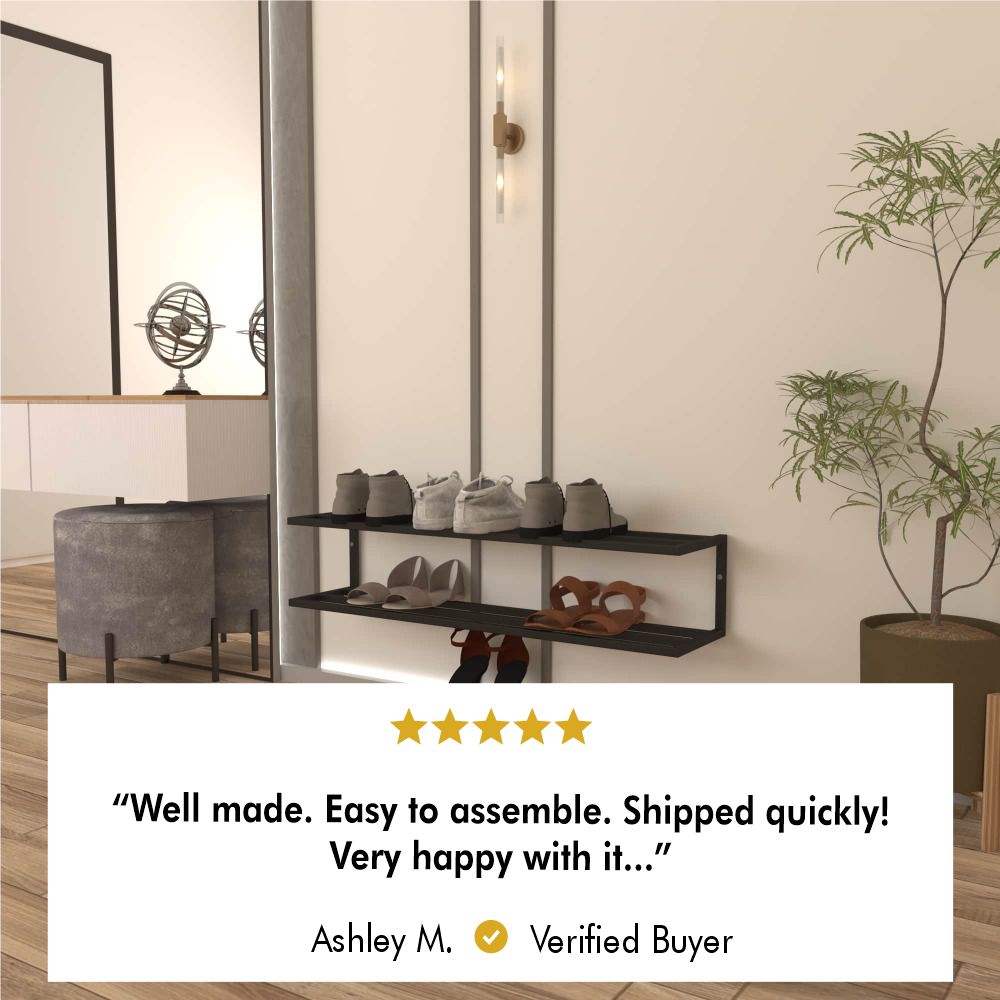 Black metal two-tier wall-mounted shoe storage shelf holding various pairs of shoes in a minimalist room with beige walls. A green potted plant stands on the right, and a stylish pendant light is mounted above the shelf. A five-star review from a verified buyer named Ashley M. appears below, stating, "Well made. Easy to assemble. Shipped quickly! Very happy with it..."