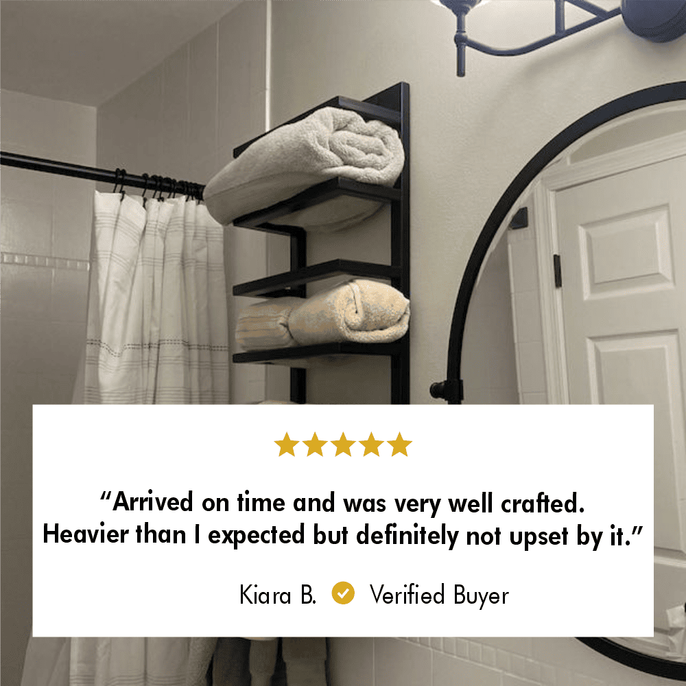 Wall-mounted black metal towel rack in a modern bathroom, holding neatly rolled towels with a round mirror and a shower curtain in the background. Review text: 'Arrived on time and was very well crafted. Heavier than I expected but definitely not upset by it.' - Kiara B., Verified Buyer.
