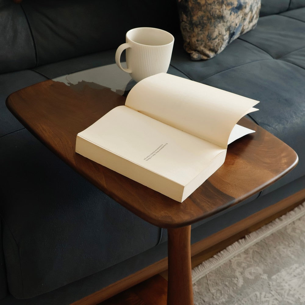 A side view of a modern walnut wood side table with a book and a coffee cup placed on top, next to a navy blue sofa.