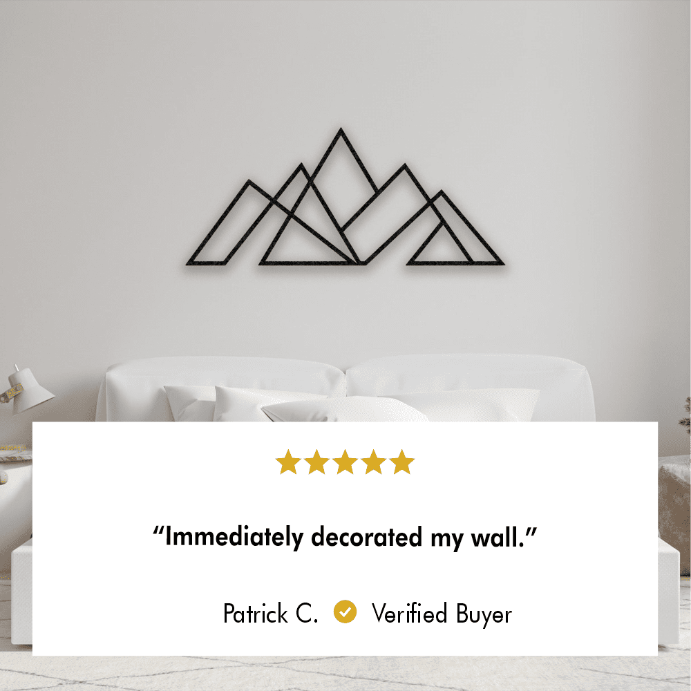 Black geometric mountain metal wall art displayed above a modern white couch in a minimalistic living room setting. The review text below reads, "Immediately decorated my wall." by Patrick C., Verified Buyer.