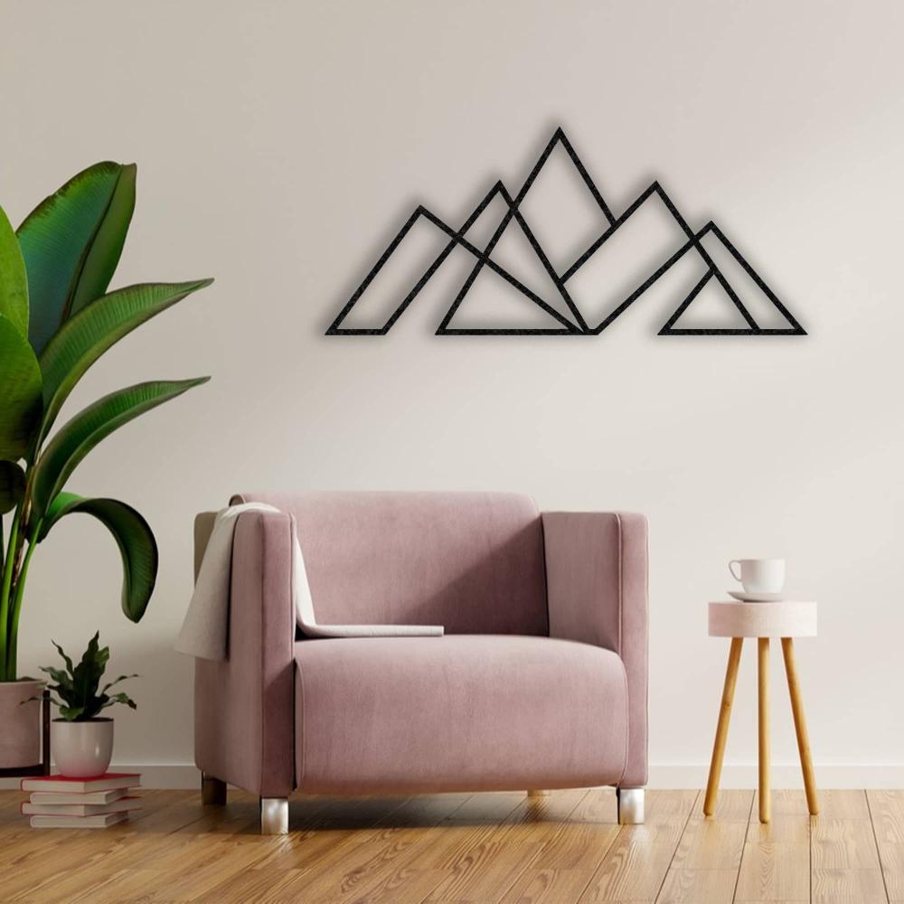 Black geometric mountain metal wall art mounted on a light-colored wall, paired with a blush pink armchair, a wooden side table with a white cup, and a vibrant green indoor plant.