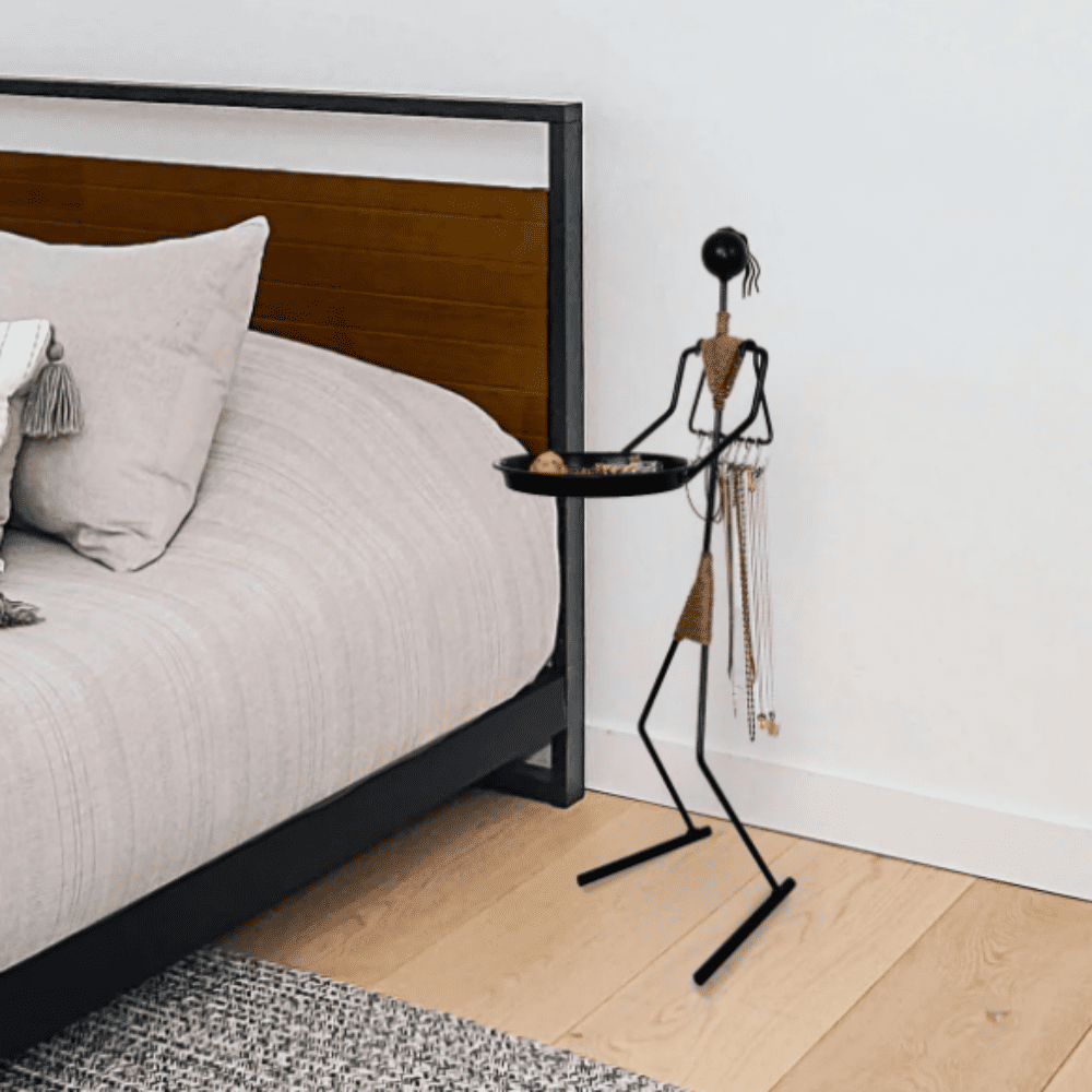 A multifunctional metal woman figure standing beside a bed, holding a black tray with jewelry and accessories, showcasing a modern and minimalist design.