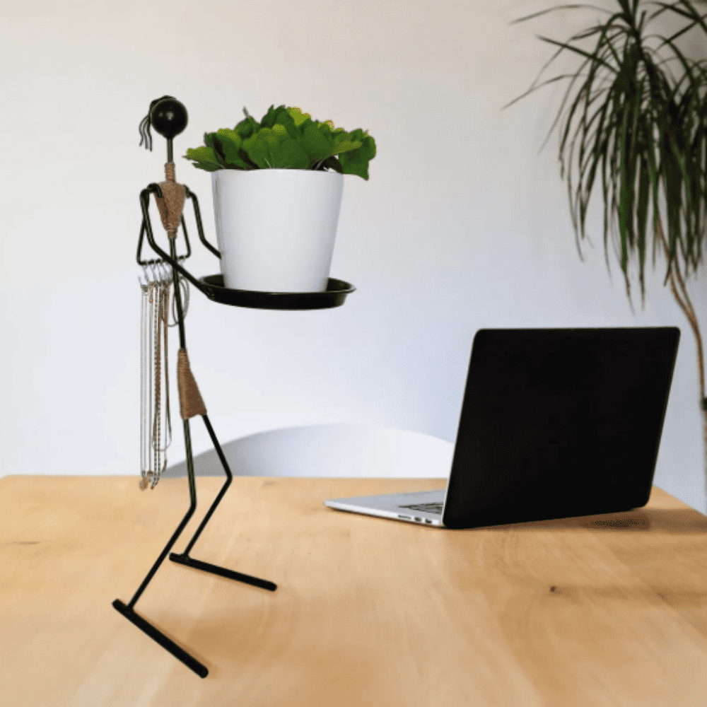 A multifunctional metal woman figure placed on a desk next to a laptop, holding a black tray with a plant and showcasing its versatility as both decor and organizer.