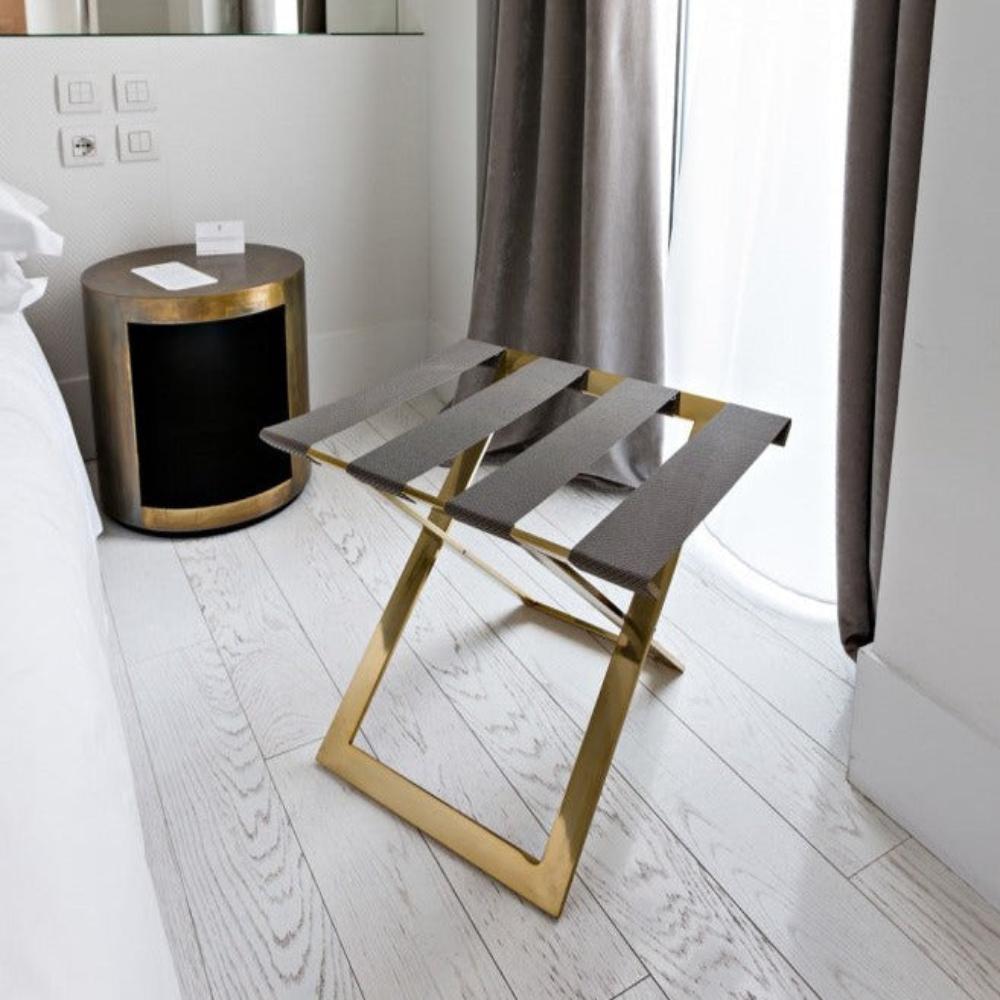 A stylish foldable metal luggage rack in gold with genuine leather straps, set near a luxurious bedside table in a bright, modern interior.