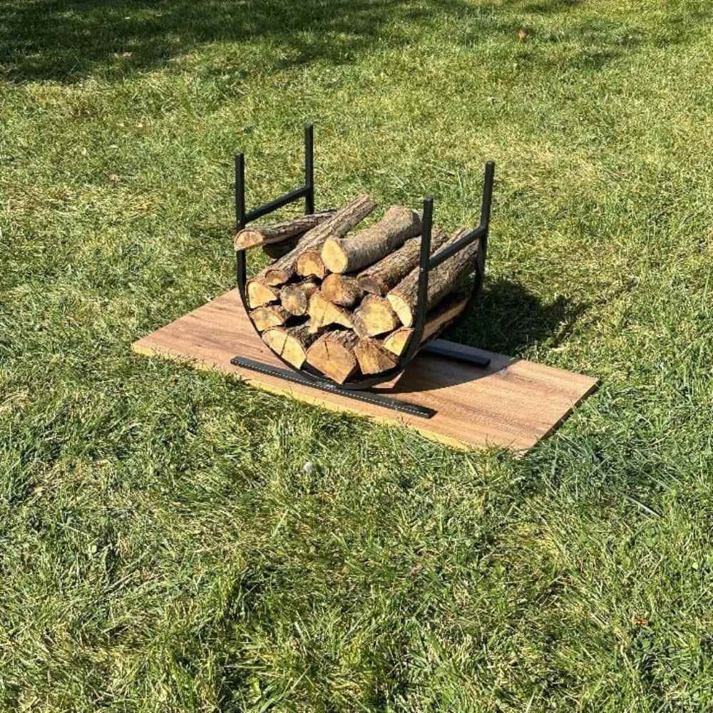 A black oval-shaped metal log holder filled with chopped logs, displayed outdoors on a wooden plank placed on a grassy field