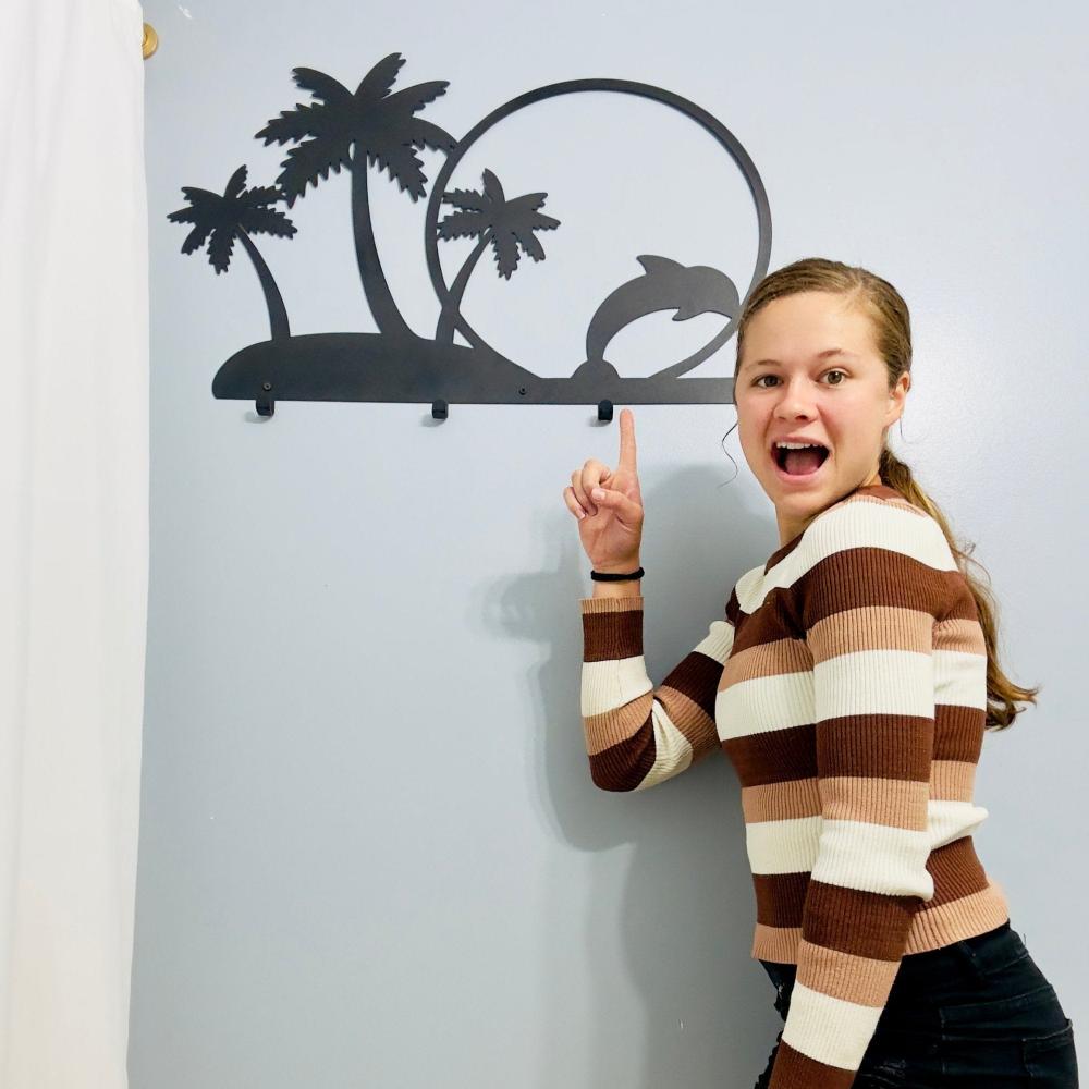 A woman enthusiastically points to a black metal wall-mounted rack featuring palm trees and a dolphin jumping through a circle. The tropical-themed design adds a fun and decorative element to the wall, with hooks for hanging items.