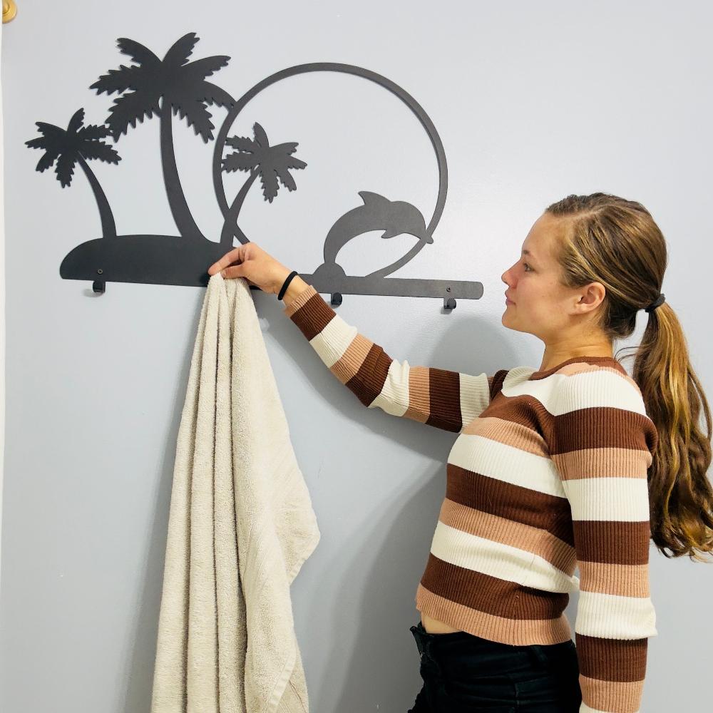 A woman hangs a towel on a black metal wall-mounted rack featuring palm trees and a dolphin jumping through a circle. The beach-themed design adds a tropical touch to the decor, offering a fun and functional hanging solution.