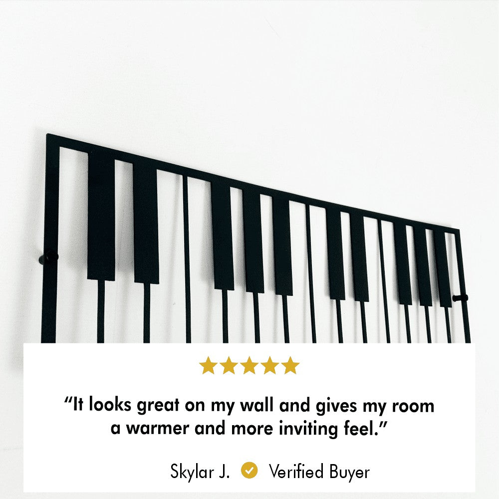 Close-up view of a metal piano keyboard wall art with a review overlay. The review reads: "It looks great on my wall and gives my room a warmer and more inviting feel." by Skylar J., Verified Buyer.