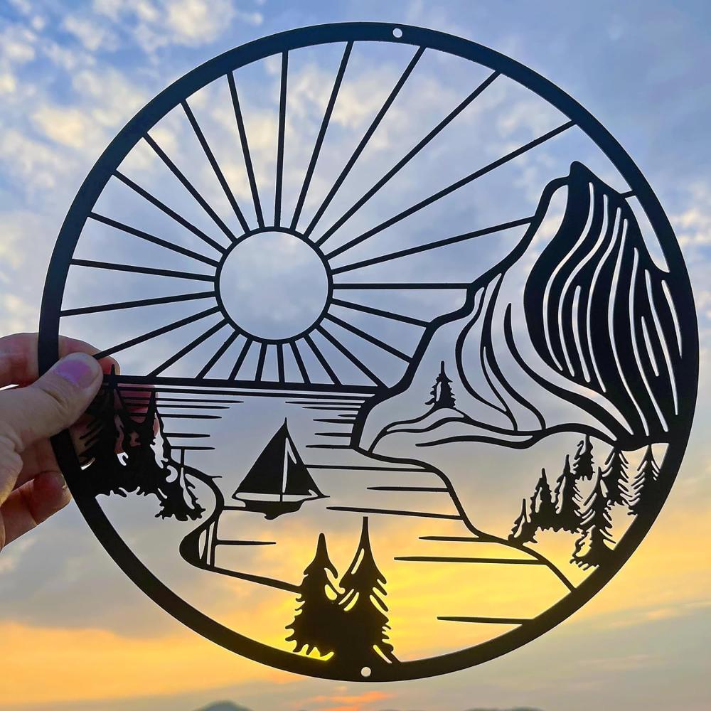 Round metal wall art depicting a serene nature scene with a sun, mountains, trees, and a sailboat, held against a vibrant sunset sky background.