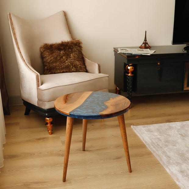 Round wooden table with blue resin accents in a living room featuring a beige armchair with a brown fur pillow and a black side table.