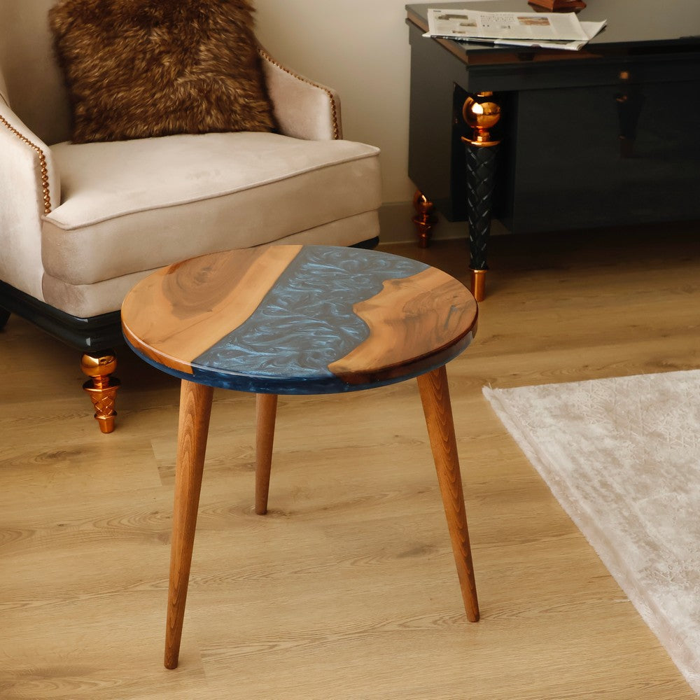 Round wooden table with blue resin accents in a living room featuring a beige armchair with a brown fur pillow and a black side table.