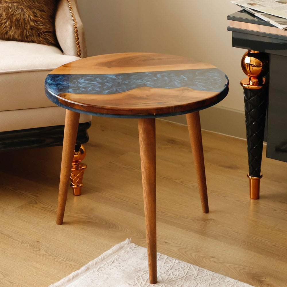 Round wooden table with blue resin accents in a living room featuring a beige armchair with a brown fur pillow and a black side table.