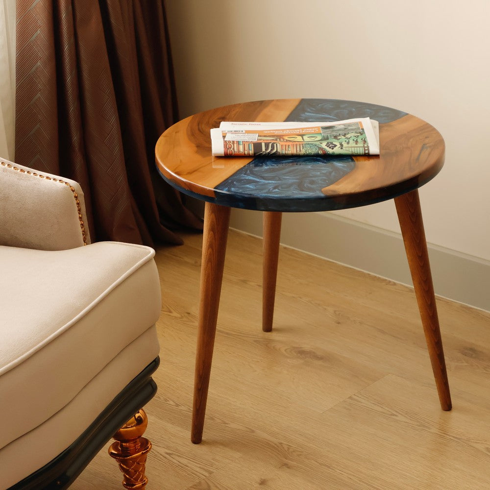 Round wooden table with blue resin accents, holding a rolled newspaper, placed next to a beige armchair in a cozy room.