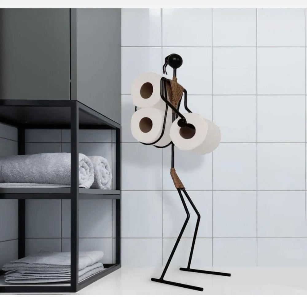 Black metal toilet paper holder in the shape of a minimalist figure, holding three rolls of toilet paper, displayed in a modern bathroom with white tiled walls and a black shelving unit containing neatly folded towels.