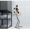 Black metal toilet paper holder in the shape of a minimalist figure, holding three rolls of toilet paper, displayed in a modern bathroom with white tiled walls and a black shelving unit containing neatly folded towels.