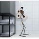 Black metal toilet paper holder in the shape of a minimalist figure, holding three rolls of toilet paper, displayed in a modern bathroom with white tiled walls and a black shelving unit containing neatly folded towels.