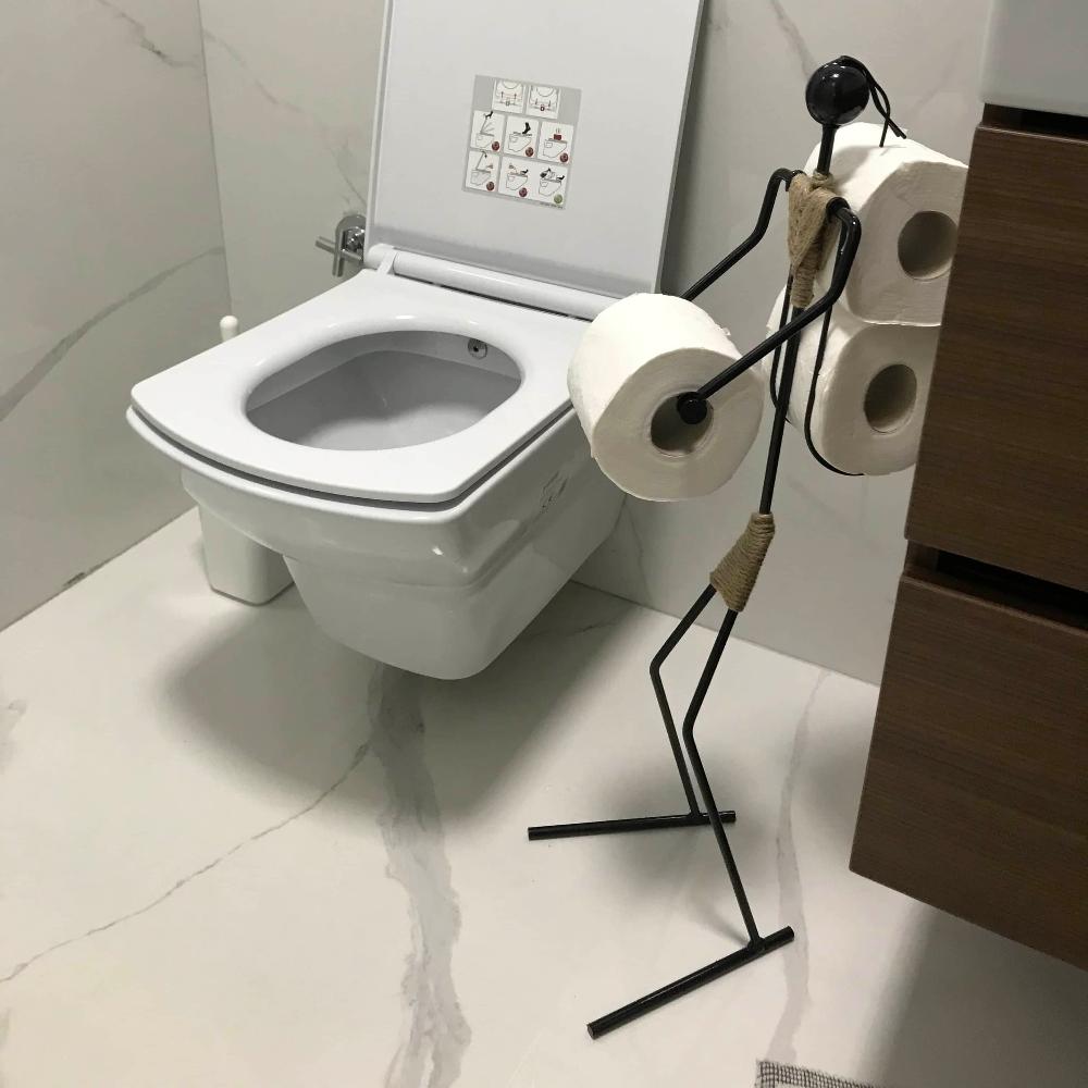 Minimalist black metal toilet paper holder shaped like a figure, holding three rolls of toilet paper, positioned next to a modern white toilet in a bathroom with marble flooring and wall tiles.