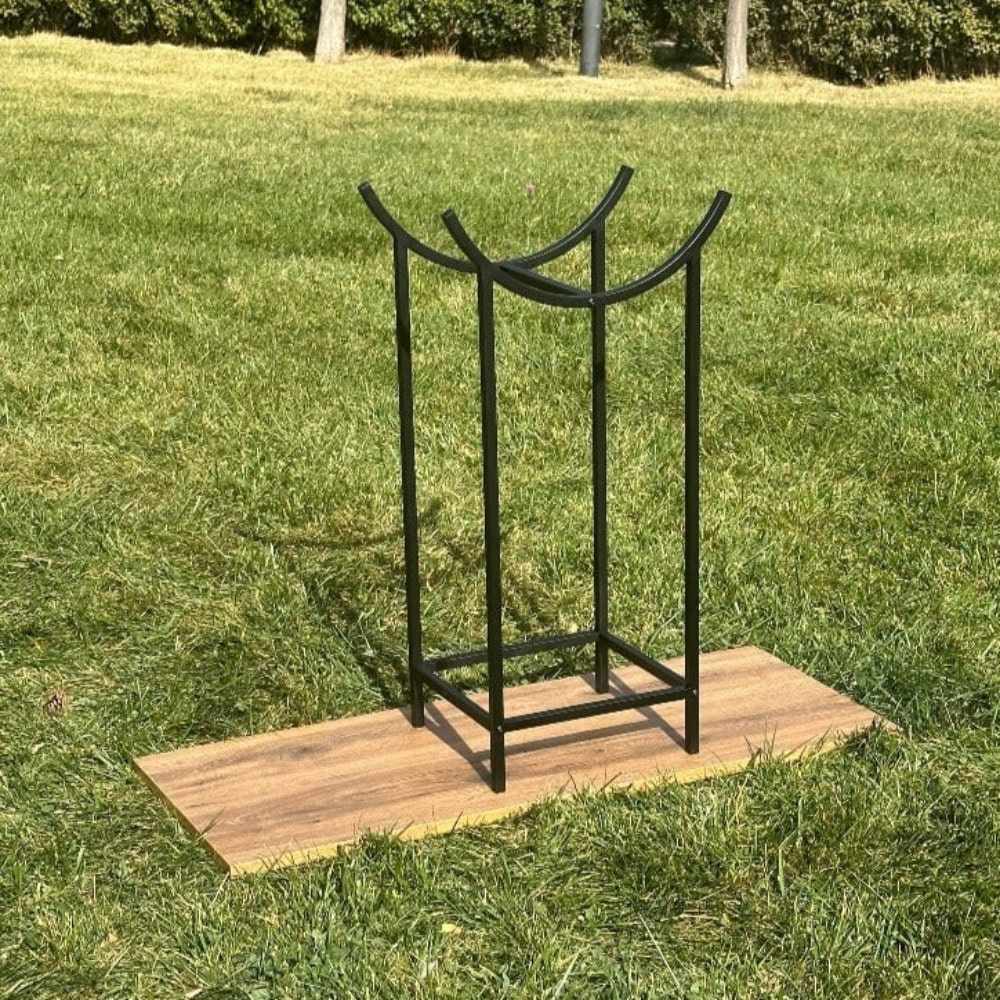 A minimalist black metal log and kindling holder with a unique curved top design placed outdoors on a wooden platform, surrounded by a lush green lawn.