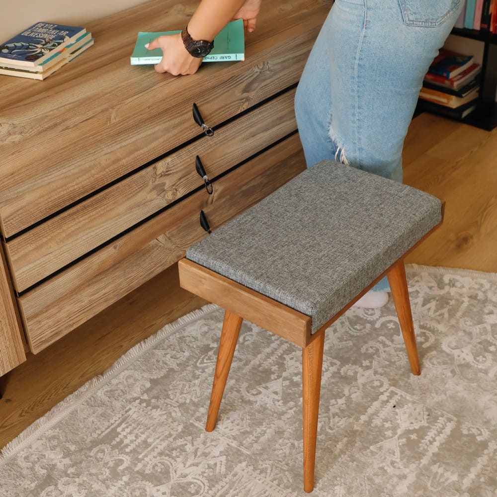 A close-up of a wooden stool with a grey cushioned seat placed on a patterned rug in front of a wooden dresser with books displayed on it.