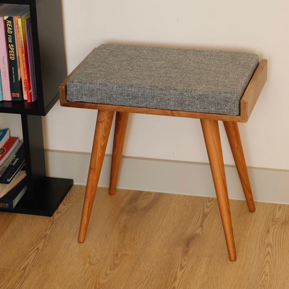 A close-up of a wooden stool with a grey cushioned seat, placed next to a black bookshelf on a wooden floor.